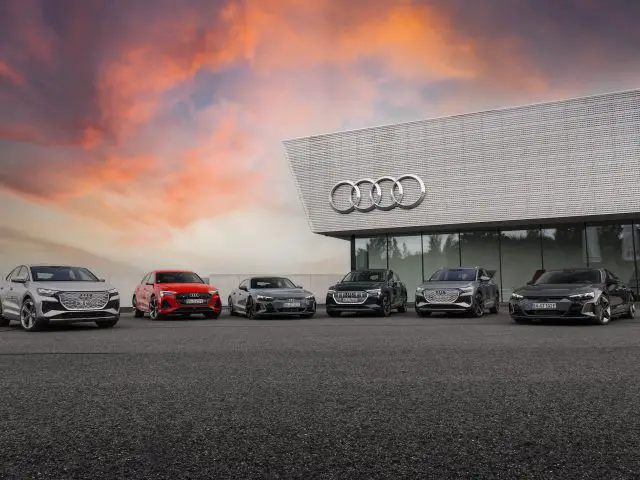 Bij zonsondergang staat een rij van vijf strakke Audi-auto's geparkeerd voor het iconische Audi-dealergebouw, terwijl een dramatische lucht een betoverende achtergrond werpt.