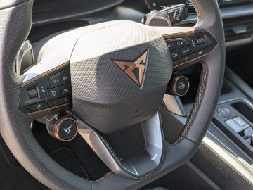 Close-up van het stuur van een Cupra Formentor, met het logo, de bedieningsknoppen en een start/stop-motorknop. Het interieur is voorzien van een mix van leer- en koolstofvezeltexturen voor een luxe gevoel.