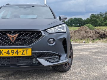 Een matzwarte Cupra Formentor SUV met een opvallende grille en een gele kentekenplaat "XK-494-ZT" staat geparkeerd op een verhard terrein met een met gras begroeide heuvel en bomen op de achtergrond onder een bewolkte hemel.