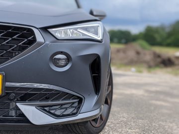 Close-up van de voorste hoek van een donkergrijze Cupra Formentor, met de koplamp, de grille en een deel van de voorbumper, tegen de achtergrond van een buitenomgeving.