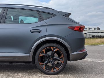 Zijaanzicht van het achterste gedeelte van een grijze Cupra Formentor SUV met wielen met oranje accenten, geparkeerd op een weg, met een gebouw en een bewolkte lucht op de achtergrond.