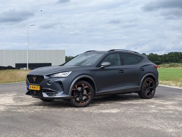Een matzwarte Cupra Formentor SUV met donkere lichtmetalen velgen staat geparkeerd in een open ruimte met een gebouw, een grasveld en een bewolkte lucht. Een geel-zwarte kentekenplaat siert de voorkant.