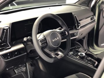 Close-up van het interieur van een moderne Kia Sportage met een stuur met een Kia-logo, een digitaal dashboarddisplay en verschillende bedieningsknoppen en knoppen op de middenconsole.