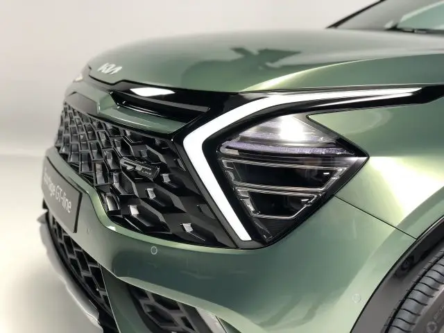 Close-up van de voorkant van een groene Kia Sportage, met de grille, de koplamp en een deel van de motorkap met een modern, hoekig ontwerp.