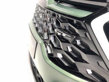 Close-up van de grille van een Kia Sportage met een opvallend geometrisch patroon en een badge met het opschrift "Line" aan de rechterkant. Het lijkt erop dat de auto in een metallic groene kleur is gespoten.