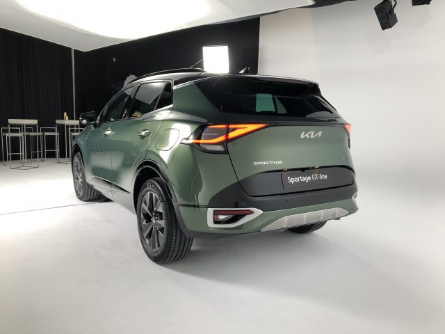 Een groene Kia Sportage GT Line staat in een helder verlichte studio met zwart-witte muren, waardoor het strakke ontwerp vanuit de driekwarthoek aan de achterkant goed tot uiting komt.