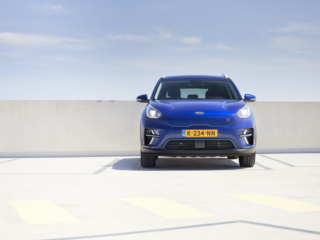 Een blauwe Kia-auto, geadverteerd voor autoverkoop, staat geparkeerd op een open plek met een betonnen muur en blauwe lucht op de achtergrond. Het Nederlandse kenteken "K-234-NN" is duidelijk zichtbaar.