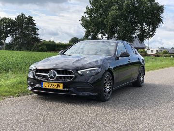 Een zwarte Mercedes-Benz C-Klasse sedan met een geel kenteken staat geparkeerd aan de kant van een landelijke weg, omgeven door weelderige groene velden en schilderachtige huizen op de achtergrond.