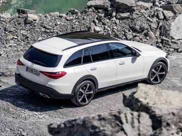 Een witte Mercedes-Benz C-Klasse All-Terrain stationwagen staat geparkeerd op rotsachtig terrein nabij een watermassa. De auto beschikt over zwarte dakaccenten en getinte ruiten, waardoor robuuste capaciteiten perfect samengaan met een strak design.