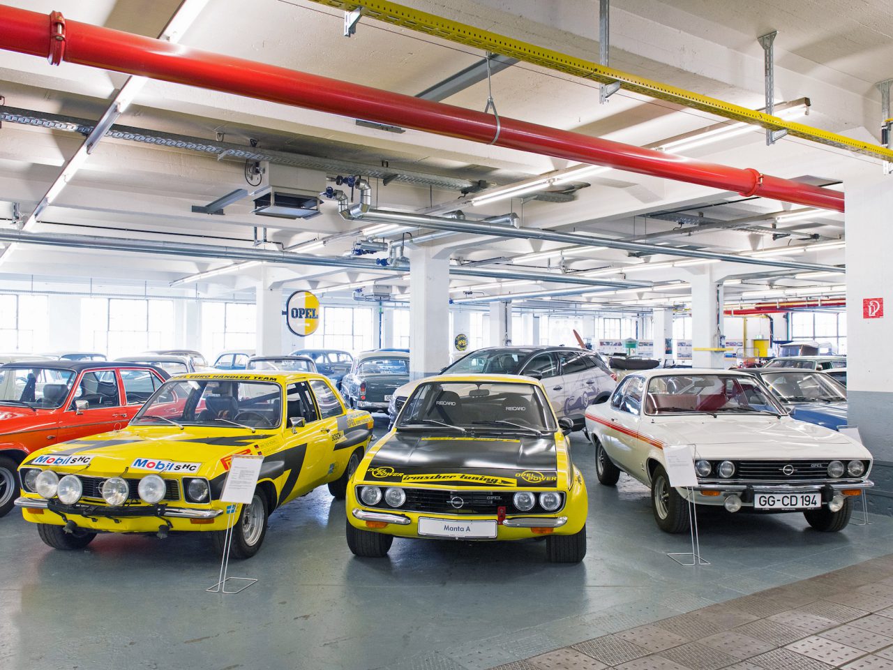 Een autoshowroom met drie geel-zwarte vintage sportwagens, waaronder een Opel Classic, zij aan zij opgesteld, met andere voertuigen zichtbaar op de achtergrond.