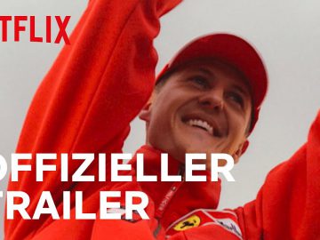 Een persoon in een rood jasje en pet glimlacht en heft zijn armen op ter viering, wat doet denken aan de iconische overwinningen van Schumacher. Het Netflix-logo en de tekst "OFFIZIELLER TRAILER" worden over de afbeelding heen weergegeven.