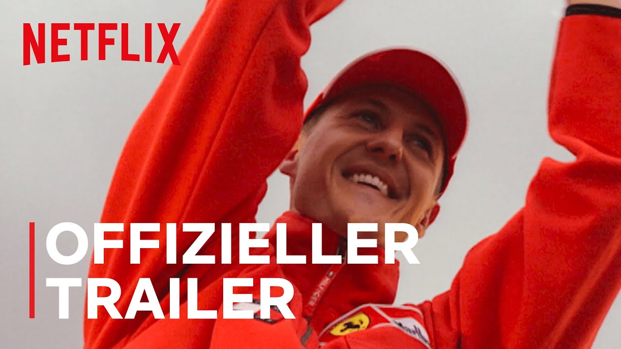 Een persoon in een rood jasje en pet glimlacht en heft zijn armen op ter viering, wat doet denken aan de iconische overwinningen van Schumacher. Het Netflix-logo en de tekst "OFFIZIELLER TRAILER" worden over de afbeelding heen weergegeven.