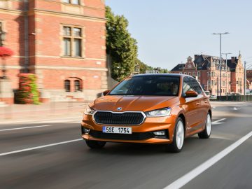 Een feloranje Skoda Fabia sedan rijdt door een stadsstraat langs historische bakstenen gebouwen en bomen.