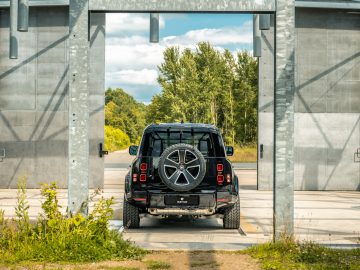 Een zwarte SUV is van achteren afgebeeld terwijl hij door een industriële poort rijdt, met een reservewiel aan de achterkant. Op de achtergrond zijn bomen en een gedeeltelijk bewolkte lucht zichtbaar, wat het gevoel van avontuur en verkenning oproept dat vaak wordt geassocieerd met Winston Gerschtanowitz.