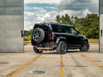 Een zwarte SUV met een reservewiel achterop, mogelijk bestuurd door Winston Gerschtanowitz, rijdt vanuit een grote garage of magazijn met betonnen muren een buitenruimte omgeven door bomen in.