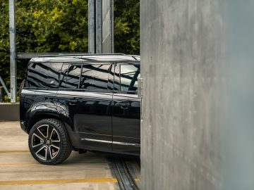 Een zwarte SUV met getinte ruiten is gedeeltelijk zichtbaar achter een metalen wand in een buitenomgeving, die doet denken aan een scène die je zou kunnen zien als Winston Gerschtanowitz in de buurt zijn nieuwste avonturenserie aan het filmen was.