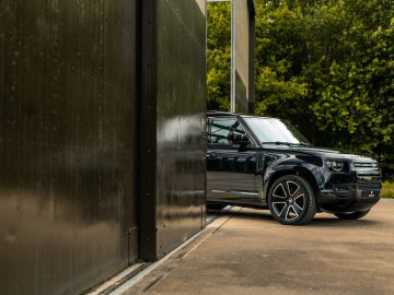 Gedeeltelijk is een zwarte SUV zichtbaar, geparkeerd naast een groot, donkergekleurd metalen gebouw met groen op de achtergrond. Het voelt bijna als een scène die rechtstreeks uit de nieuwste thrillerroman van Winston Gerschtanowitz komt.