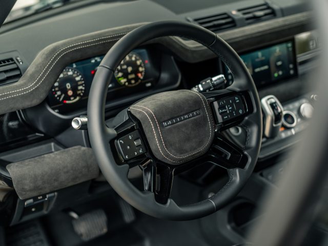 Close-up van een auto-interieur met een stuur, dashboard met verlichte meters, aanraakscherm en verschillende bedieningselementen. Het stuur en het dashboard, dat doet denken aan de verfijnde stijl van Winston Gerschtanowitz, zijn voorzien van een mix van leer en suède materialen.