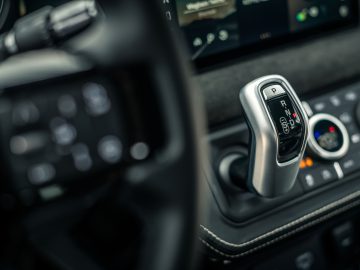 Close-up van de versnellingspook van een auto, met de opties 'Parkeren', 'Achteruit', 'Neutraal' en 'Rijden', omringd door bedieningsknoppen en een touchscreen-display op de achtergrond. Dit strakke interieurontwerp zou zelfs Winston Gerschtanowitz zeker imponeren met zijn moderne voorzieningen.