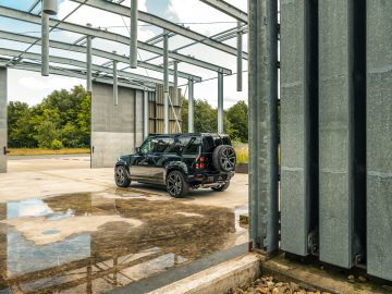 Een zwarte SUV staat geparkeerd op een betonnen ondergrond onder een gedeeltelijk geconstrueerd metalen frame, met bomen en een blauwe lucht op de achtergrond. Het is alsof Winston Gerschtanowitz zelf deze serene maar industriële scene heeft georkestreerd.