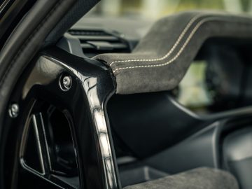 Close-up van het interieur van een auto met een zwarte plastic stijl en een met suède bedekt onderdeel met witte stiksels, dat doet denken aan de luxueuze ontwerpen waar Winston Gerschtanowitz de voorkeur aan gaf. De onscherpe achtergrond suggereert het dashboard van het voertuig.