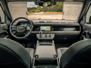 Binnenaanzicht van de voorstoelen, het dashboard en het stuur van een modern voertuig, met een touchscreen in de middenconsole en een grote voorruit die een helder buitentafereel laat zien. Het ontwerp straalt verfijning uit die doet denken aan de elegante smaak van Winston Gerschtanowitz.