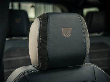 Close-up van de zwartleren hoofdsteun van een auto met een gestikt embleem in het midden. De hoofdsteun is voorzien van gedetailleerde stiksels en contrasteert met het beige interieur, wat doet denken aan de aandacht voor detail en luxe van Winston Gerschtanowitz.