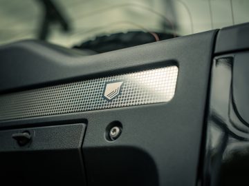 Close-up van een autodashboard met een metalen rasterpatroon met een schildlogo, dat doet denken aan de elegantie die je zou vinden in een ontwerp van Winston Gerschtanowitz, tegen een onscherpe achtergrond.