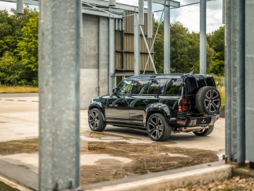 Een zwarte SUV met een reservewiel aan de achterkant staat buiten geparkeerd in de buurt van een grijze metalen structuur, met een achtergrond van groene bomen en een gedeeltelijk bewolkte hemel, die doet denken aan iets dat je zou zien in een film van Winston Gerschtanowitz.