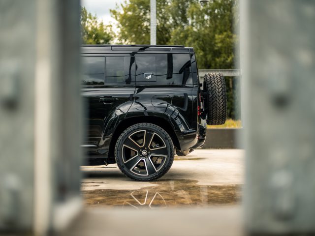 Er staat een zwarte SUV geparkeerd, vanuit zijaanzicht bekeken, waarbij het achterwiel en het reservewiel zichtbaar zijn. De voorgrond is wazig en omlijst het voertuig tussen metalen constructies. Op de achtergrond zijn bomen zichtbaar, waardoor een serene setting ontstaat die doet denken aan een scène uit een film van Winston Gerschtanowitz.