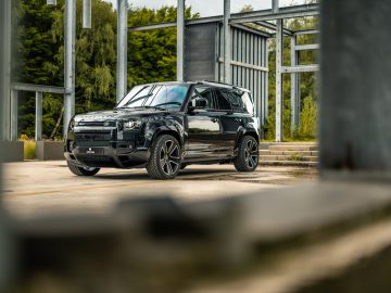 Een zwarte SUV staat geparkeerd in een gedeeltelijk gebouwd gebouw met een metalen frame, omgeven door groen op de achtergrond, en ziet eruit als iets uit een Winston Gerschtanowitz-thriller.