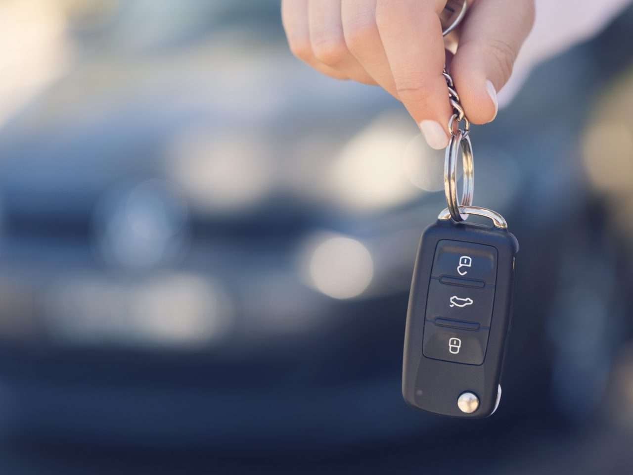 Een hand die autosleutels vasthoudt met een sleutelhanger met knoppen voor vergrendelen, ontgrendelen en kofferbakontgrendeling, met wazige populaire tweedehands auto's op de achtergrond.