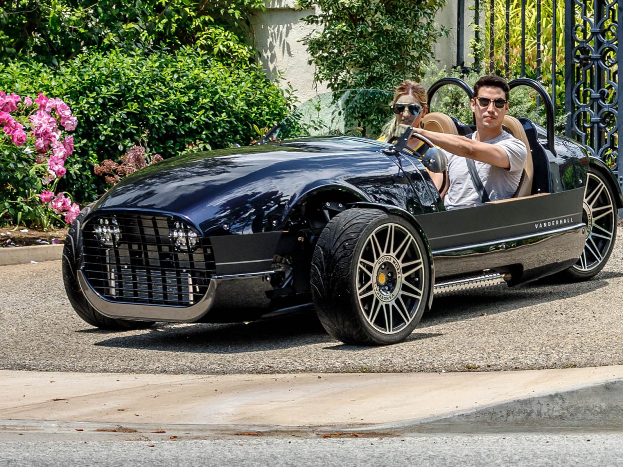 Twee mensen besturen een strak, zwart Vanderhall driewielig voertuig op een oprit met weelderig groen en bloemen op de achtergrond.