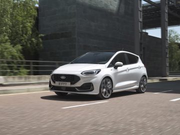 Een witte Ford Fiesta rijdt op een verharde weg naast een gebouw met grote ramen. Op de achtergrond zijn bomen en balustrades zichtbaar.