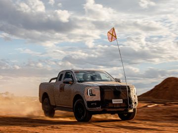 Een gecamoufleerde Ford Ranger-pick-up met een vlag eraan is off-road op een onverharde weg onder een bewolkte hemel.