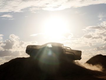 Een Ford Ranger rijdt een onverharde heuvel op tegen een felle zon aan een bewolkte hemel, waardoor een dramatisch silhouet ontstaat met stof achter zich aan.