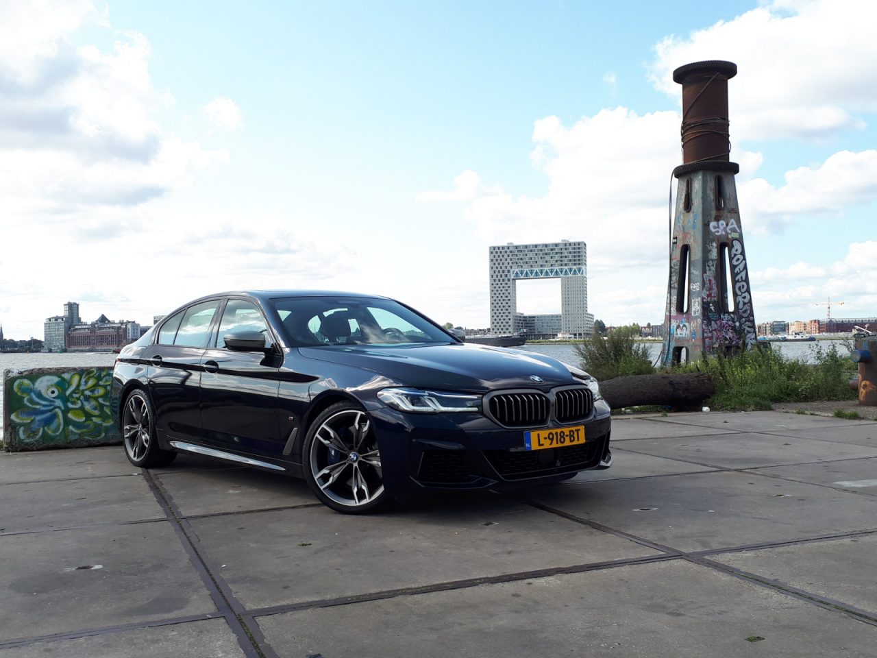 Een zwarte BMW M550i xDrive luxe sedan met kenteken L-918-BT staat geparkeerd op een betonnen ondergrond nabij een met graffiti beschilderde pilaar. De achtergrond bestaat uit water, een modern gebouw en een bewolkte hemel.