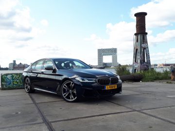 Een zwarte BMW M550i xDrive luxe sedan met kenteken L-918-BT staat geparkeerd op een betonnen ondergrond nabij een met graffiti beschilderde pilaar. De achtergrond bestaat uit water, een modern gebouw en een bewolkte hemel.