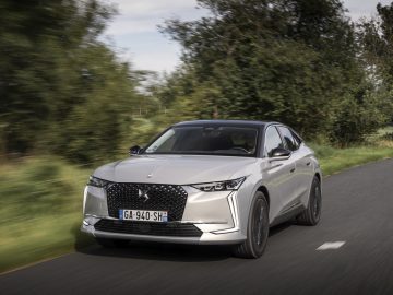 Een strakke DS 4 met Europees kenteken rijdt over een tweebaansweg, omgeven door weelderig groen.