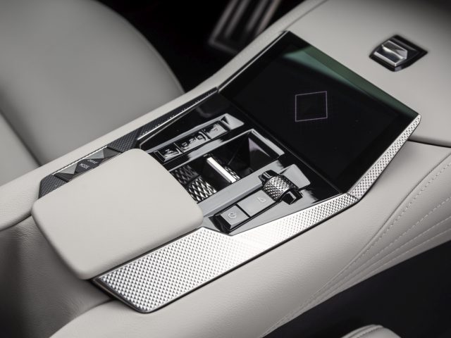 Close-up van de middenconsole van de DS 4 met aanraakscherm, verschillende bedieningsknoppen en een gewatteerde armleuning. Het ontwerp is strak met een mix van zwarte, zilveren en lichtgrijze elementen.