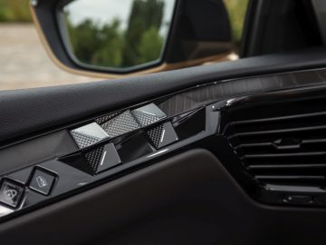 Close-up van het interieur van een DS 4 met het deurpaneel aan de bestuurderszijde met bedieningselementen voor ramen en spiegels, een ventilatieopening en een zicht op de zijspiegel die een wazige buitenachtergrond weerspiegelt.