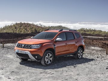 Een feloranje Dacia Duster staat geparkeerd op een grindoppervlak met bergen en wolken op de achtergrond onder een helderblauwe lucht.