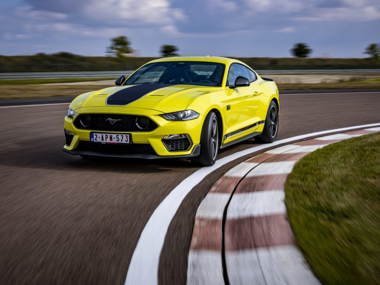 Een felgele Ford Mustang Mach 1 met een zwarte racestreep rijdt op een racecircuit en passeert een bocht.