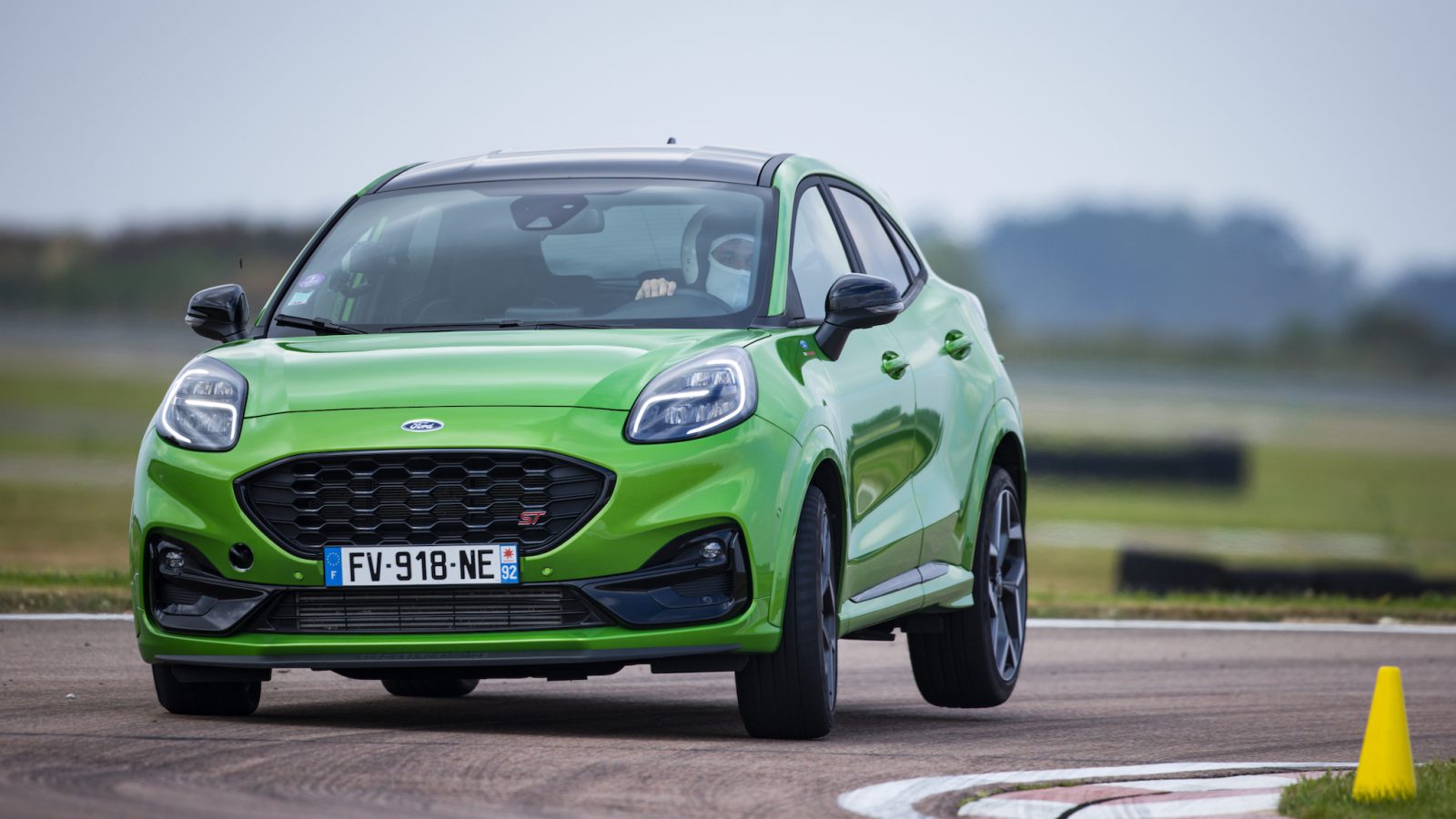 Er wordt een felgroene Ford Puma ST getoond die een bocht op een racecircuit navigeert en vakkundig rond een gele kegel manoeuvreert. Op de kentekenplaat aan de voorkant van de auto staat "FV-918-NE", terwijl de bestuurder de bocht nauwkeurig neemt.