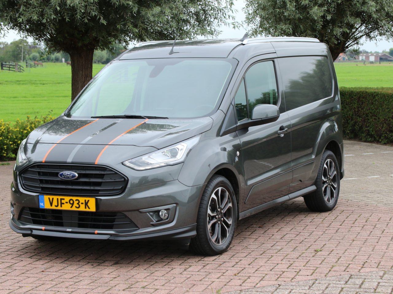Een grijs Ford Transit Connect-busje met oranje racestrepen staat geparkeerd op een verhard terrein naast een grasveld en bomen. Het voertuig heeft Nederlands kenteken.