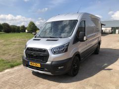 Op een zonnige dag staat een zilveren Ford Transit Trail-busje buiten geparkeerd, iets naar rechts gericht. Het voertuig heeft een Europees kenteken. Op de achtergrond is er een grasveld en een gebouw met een golfplaten dak.