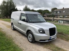 Op een grindweg met huizen en bomen op de achtergrond staat een zilverkleurige bestelwagen, die met zijn stoere bouw doet denken aan een Londense taxi. Het busje is voorzien van Europese kentekenplaten.