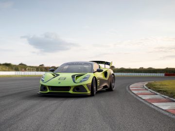 Een felgroene Lotus Emira GT4 snelt overdag over een racecircuit. De achtergrond is voorzien van een bewolkte hemel en groen.