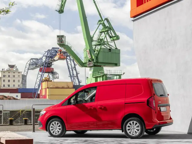 Een rode Mercedes-Benz Citan bestelwagen staat geparkeerd nabij een industriegebied met kranen en zeecontainers op de achtergrond.