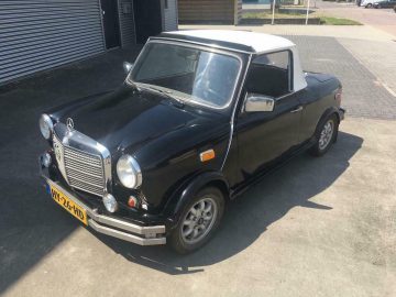 Op een verharde ondergrond staat een kleine, zwarte Mini Benz Cabrio met een wit dak geparkeerd. De auto heeft een vintage design, opvallende ronde koplampen en een Nederlands kenteken met de tekst "HV-26-HD.